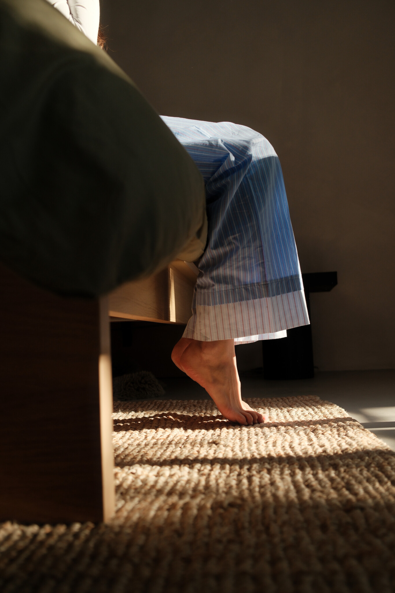 Person sitting on the edge of a bed in early morning light, bare feet resting on a woven rug