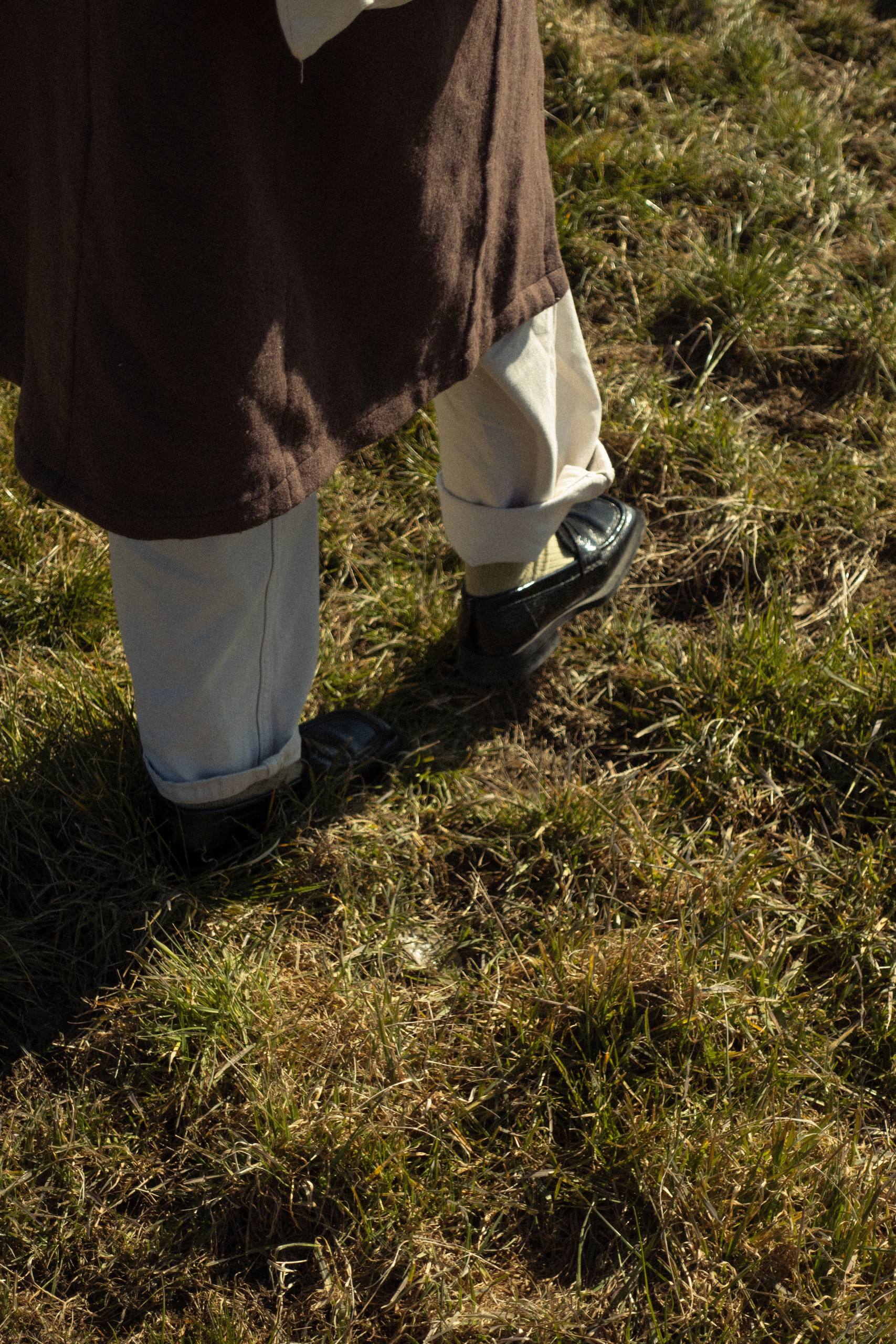 Someone walking across sunlit grass in a brown coat and cream trousers