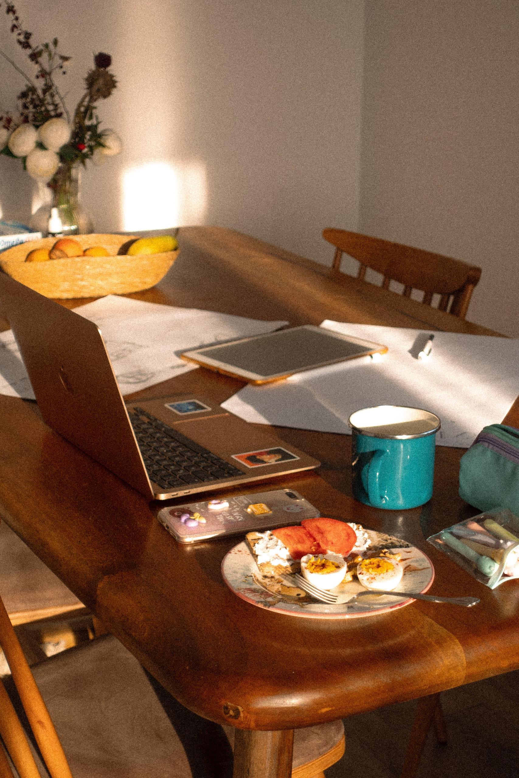 A laptop, a mug and a half-eaten breakfast on a sunlit kitchen table at home