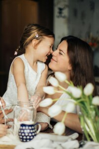 A mother and her young daughter touching noses and laughing at a kitchen table, white tulips in the foreground