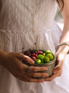 A woman in a white eyelet dress holds a ceramic bowl of red cherries and green plums close to her body