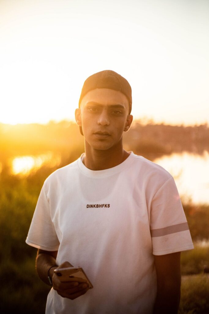 A man standing outdoors in low evening sun, holding his phone, looking straight ahead with a neutral expression